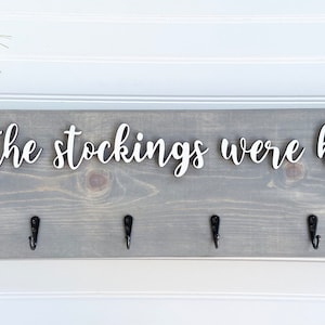May include: A gray wooden sign with the text "and the stockings were hung..." in white script. The sign has six black hooks for hanging stockings. Greenery and red beads are in the upper left corner.