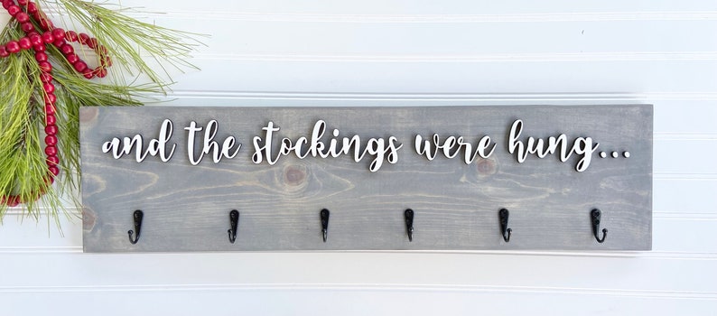 May include: A gray wooden sign with the text "and the stockings were hung..." in white script. The sign has six black hooks for hanging stockings. Greenery and red beads are in the upper left corner.