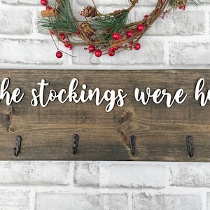 May include: A rustic wooden sign with the phrase "and the stockings were hung..." in white script. The sign has six black metal hooks for hanging stockings. A festive wreath with red berries and greenery is above the sign.