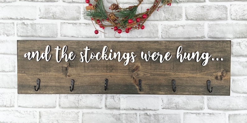 May include: A rustic wooden sign with the phrase "and the stockings were hung..." in white script. The sign has six black metal hooks for hanging stockings. A festive wreath with red berries and greenery is above the sign.