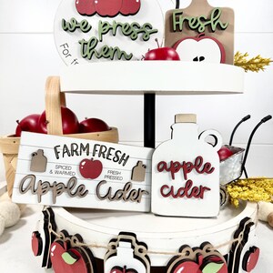 Op de afbeelding: Een wit dienblad met twee niveaus en houten decoraties in appelthema. Items zijn onder meer bordjes met de teksten U-Pick them we press them, Farm Fresh Apple Cider en apple cider, samen met een slinger van kleine houten appelvormen.