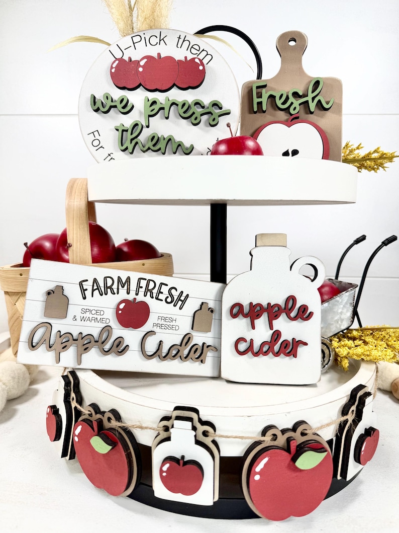Op de afbeelding: Een wit dienblad met twee niveaus en houten decoraties in appelthema. Items zijn onder meer bordjes met de teksten U-Pick them we press them, Farm Fresh Apple Cider en apple cider, samen met een slinger van kleine houten appelvormen.