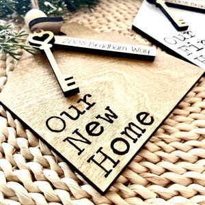 May include: Wooden ornament shaped like a house with a keyhole and a golden key. The ornament is engraved with the text "Our New Home" and the address "2008 Bradham Way".