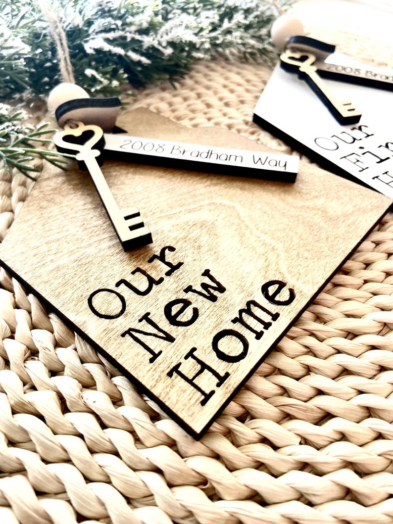 May include: Wooden ornament shaped like a house with a keyhole and a golden key. The ornament is engraved with the text "Our New Home" and the address "2008 Bradham Way".