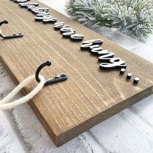 May include: A wooden sign with the phrase "The stockings were hung..." in white and black lettering. The sign has three black metal hooks and a white rope. The wood is stained a light brown color.