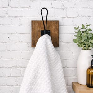 May include: A white textured towel hangs from a black metal loop hook mounted on a square, dark-stained wooden base. The hook is secured with visible screws, providing a sturdy support for the fabric while maintaining a clean, functional design.