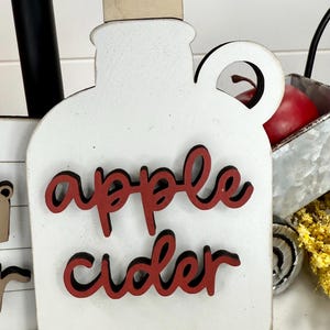 Op de afbeelding: Een witte houten uitsparing in de vorm van een kan met een handvat en een lichtbruine stop. De tekst apple cider is in rode letters met een zwarte omlijning op de voorzijde aangebracht. Het object heeft een platte voet en een ronde bovenkant.
