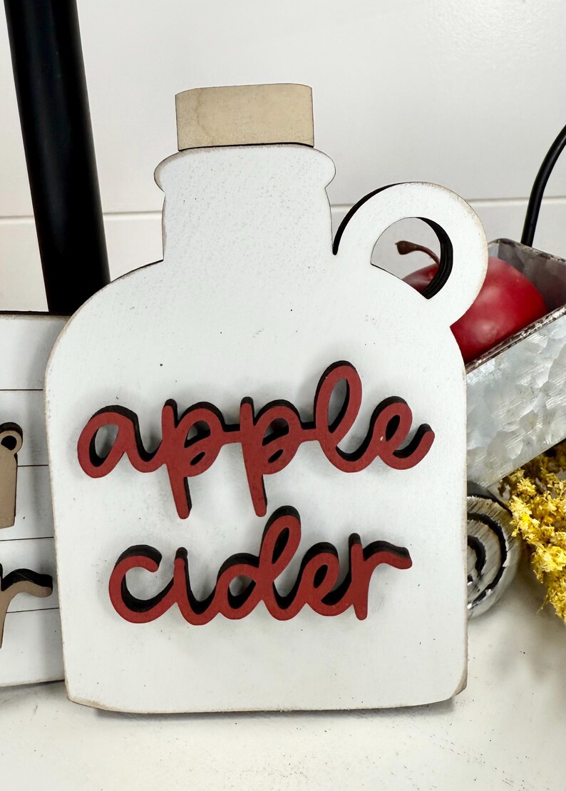 Op de afbeelding: Een witte houten uitsparing in de vorm van een kan met een handvat en een lichtbruine stop. De tekst apple cider is in rode letters met een zwarte omlijning op de voorzijde aangebracht. Het object heeft een platte voet en een ronde bovenkant.