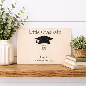 May include: A wooden graduation photo frame with the words "Little Graduate" and a black mortarboard. The frame includes space for a photo and the name "Lincoln" with "Kindergarten 2025" below. The frame is displayed on a wooden shelf.