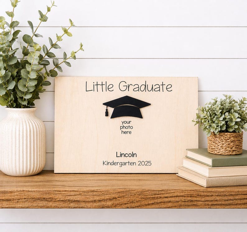 May include: A wooden graduation photo frame with the words "Little Graduate" and a black mortarboard. The frame includes space for a photo and the name "Lincoln" with "Kindergarten 2025" below. The frame is displayed on a wooden shelf.