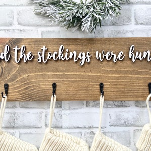May include: Wooden Christmas stocking holder with the phrase "and the stockings were hung..." in white script. The brown wooden board has four black hooks, each holding a cream-colored stocking. A sprig of frosted greenery sits above the board.