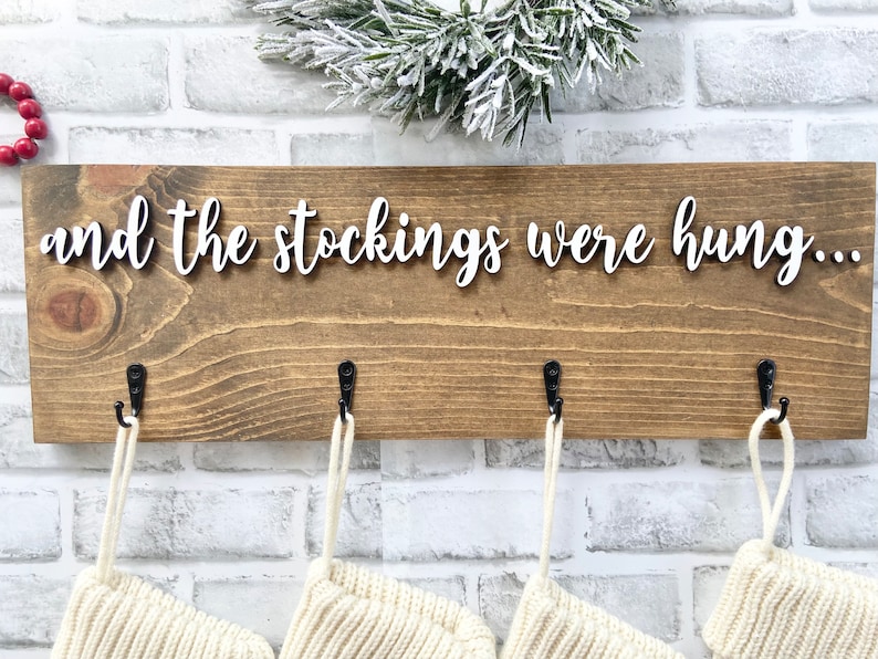 May include: Wooden Christmas stocking holder with the phrase "and the stockings were hung..." in white script. The brown wooden board has four black hooks, each holding a cream-colored stocking. A sprig of frosted greenery sits above the board.