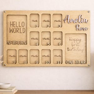 Może przedstawiać: Drewniana tablica na zdjęcia z miejscami na zdjęcia od urodzenia do roku. Tablica zawiera imię "Amelia Rose" oraz napisy "Hello World", "Newborn" i "Happy first Birthday!"