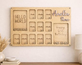 Puede incluir: Tablero de fotos de madera con espacios para fotos desde el nacimiento hasta el primer año. El tablero incluye el nombre "Amelia Rose" y las frases "Hello World", "Newborn" y "Happy first Birthday!"