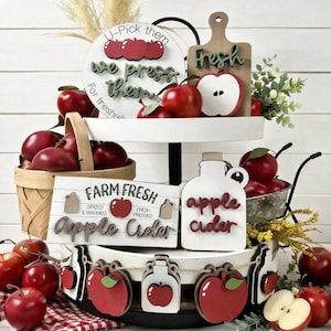 Op de afbeelding: Een wit dienblad met twee niveaus, versierd met houten bordjes met appelthema, waaronder een rond bordje met de tekst U-Pick them we press them, een snijplank met Fresh en een rechthoekig bordje met FARM FRESH Apple Cider.