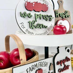 Op de afbeelding: Een etag&egrave;re met ronde en rechthoekige houten bordjes met appel- en ciderthema's. De bordjes bevatten teksten zoals U-Pick them, we press them, For fresh pressed cider, Farm Fresh en apple cider, geaccentueerd met rode appel-uitsparingen.