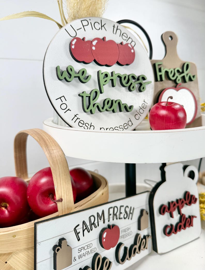 Op de afbeelding: Een etag&egrave;re met ronde en rechthoekige houten bordjes met appel- en ciderthema's. De bordjes bevatten teksten zoals U-Pick them, we press them, For fresh pressed cider, Farm Fresh en apple cider, geaccentueerd met rode appel-uitsparingen.