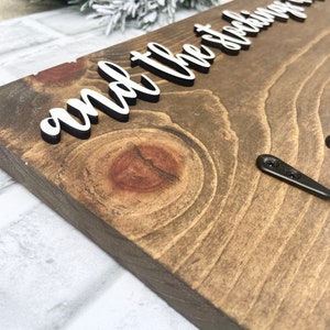 May include: Wooden sign with the phrase "and the stockings were hung" in white, raised lettering. The sign is made of stained wood with visible grain and a dark brown finish. Two black metal hooks are attached to the bottom right corner.