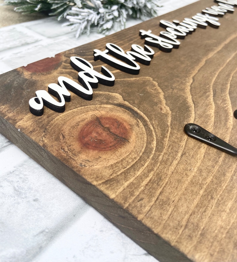 May include: Wooden sign with the phrase "and the stockings were hung" in white, raised lettering. The sign is made of stained wood with visible grain and a dark brown finish. Two black metal hooks are attached to the bottom right corner.