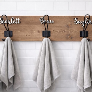 May include: A wooden wall-mounted towel rack with three black metal hooks. Each hook holds a gray towel. The names "Scarlett", "Beau", and "Saylor" are displayed above each hook in a cursive font.