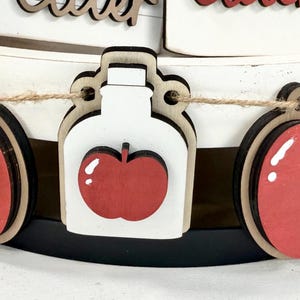 Op de afbeelding: Een decoratieve slinger met afwisselende houten uitsparingen van rode appels met groene blaadjes en witte potten met rode appelaccenten, allemaal aan elkaar geregen met een dun bruin touwtje op een wit en zwart etag&egrave;replateau.