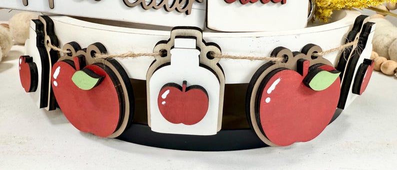Op de afbeelding: Een decoratieve slinger met afwisselende houten uitsparingen van rode appels met groene blaadjes en witte potten met rode appelaccenten, allemaal aan elkaar geregen met een dun bruin touwtje op een wit en zwart etag&egrave;replateau.
