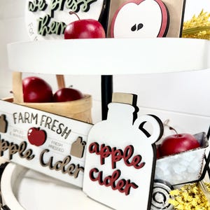 Op de afbeelding: Een etag&egrave;re met houten bordjes voorzien van appelthema teksten en afbeeldingen. De bordjes bevatten de teksten U-Pick them, we press them, Fresh, Farm Fresh en apple cider. Rode appelornamenten en een klein mandje zijn ook aanwezig.