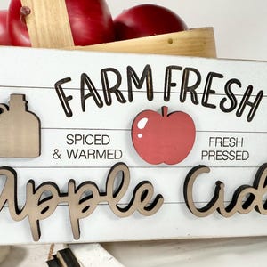Op de afbeelding: Een rechthoekig wit houten bord met horizontale lijnen met de zwarte tekst FARM FRESH en Apple Cider in cursief schrift. Het bevat een rode appel-uitsparing, twee beige kruik-uitsparingen en de zwarte tekst SPICED & WARMED en FRESH PRESSED.