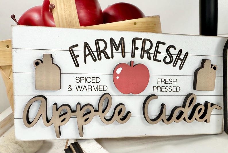 Op de afbeelding: Een rechthoekig wit houten bord met horizontale lijnen met de zwarte tekst FARM FRESH en Apple Cider in cursief schrift. Het bevat een rode appel-uitsparing, twee beige kruik-uitsparingen en de zwarte tekst SPICED & WARMED en FRESH PRESSED.