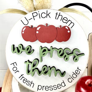 Op de afbeelding: Een rond wit houten bord met drie rode appeluitsparingen aan de bovenkant. De tekst luidt U-Pick them in het zwart, we press them in verhoogd groen schrift, en For fresh pressed cider in het zwart. Een rode appel ligt rechtsonder.