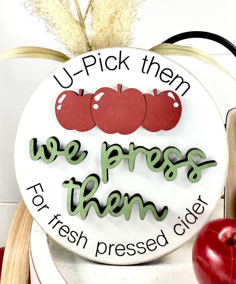 Op de afbeelding: Een rond wit houten bord met drie rode appeluitsparingen aan de bovenkant. De tekst luidt U-Pick them in het zwart, we press them in verhoogd groen schrift, en For fresh pressed cider in het zwart. Een rode appel ligt rechtsonder.