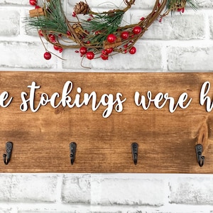 May include: Wooden sign with the phrase "and the stockings were hung..." in white script. The sign is brown with six black hooks for hanging stockings. A wreath with red berries and greenery is above the sign.