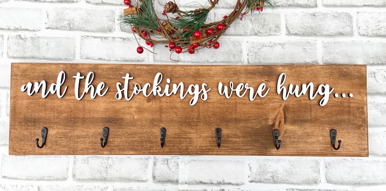 May include: Wooden sign with the phrase "and the stockings were hung..." in white script. The sign is brown with six black hooks for hanging stockings. A wreath with red berries and greenery is above the sign.
