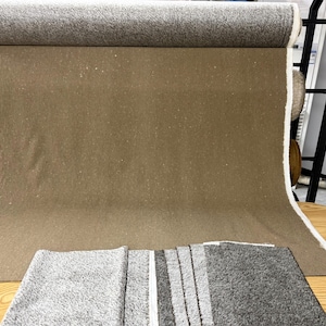 May include: A large roll of brown fabric with a textured surface, partially unrolled to reveal the material. A folded gray and white striped rug is in the foreground. The fabric is likely for upholstery or home decor.