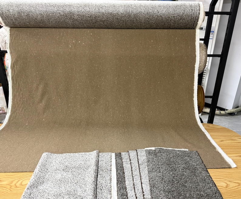 May include: A large roll of brown fabric with a textured surface, partially unrolled to reveal the material. A folded gray and white striped rug is in the foreground. The fabric is likely for upholstery or home decor.