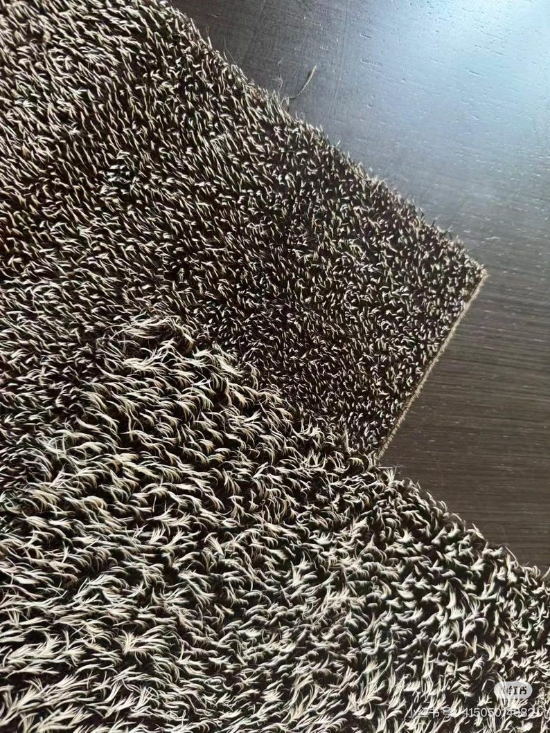 May include: Close-up of a textured, brown and white rug. The rug has a shaggy appearance with a dense pile of fibers. The fibers are a mix of brown and white, creating a speckled effect. The rug is on a dark brown surface.