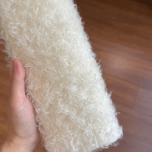 Può includere: Un tessuto rettangolare bianco sporco con una trama densa e riccia, simile alla lana di pecora. Il tessuto è tenuto in una mano, mostrando il suo aspetto morbido e soffice. Lo sfondo è una superficie di legno.