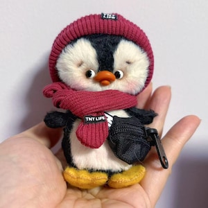 Puede incluir: Un pequeño pingüino de peluche que lleva un gorro y una bufanda de punto rojo con el texto "TNT LIFE" en la bufanda. El pingüino lleva una pequeña bolsa negra con cremallera.