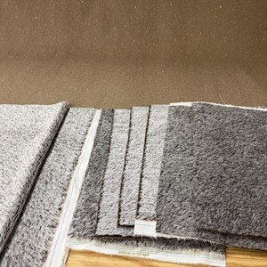 May include: A collection of gray and charcoal-colored fabric swatches. The fabric has a textured, speckled appearance. The swatches are arranged in a layered display, showcasing variations in color and texture. The background is a solid brown.