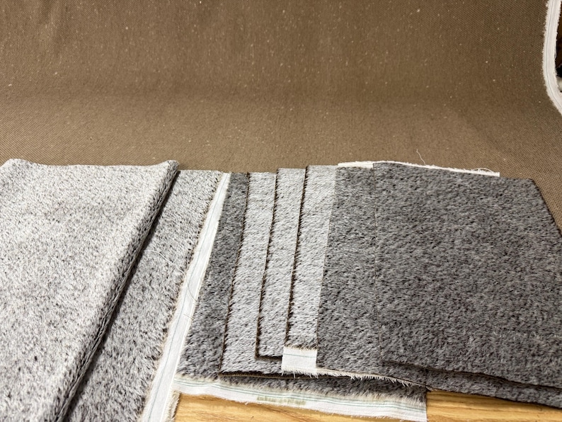 May include: A collection of gray and charcoal-colored fabric swatches. The fabric has a textured, speckled appearance. The swatches are arranged in a layered display, showcasing variations in color and texture. The background is a solid brown.