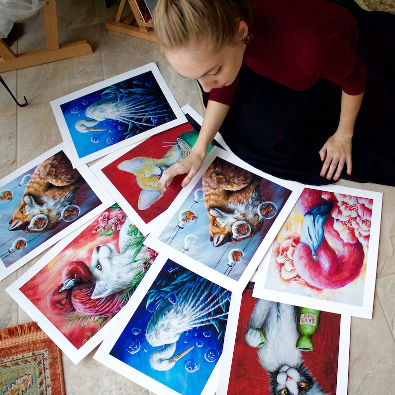 K&ouml;nnte beinhalten: Eine Sammlung von 10 Kunstdrucken mit verschiedenen Tierillustrationen. Die Drucke werden auf einem Tisch ausgestellt, wobei eine Hand auf einen der Drucke zeigt. Die Drucke zeigen eine Vielzahl von Tieren, darunter einen Flamingo, eine Katze, einen Fuchs, einen Vogel und eine Katze.