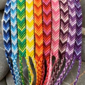 May include: A collection of colourful chevron friendship bracelets in various shades of blue, green, yellow, orange, pink, and purple.