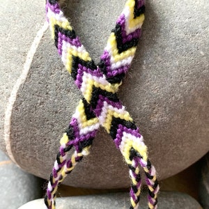 May include: A friendship bracelet with a chevron pattern in purple, yellow, white and black colours.