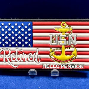 Puede incluir: Un parche rectangular con el diseño de la bandera estadounidense, las palabras "Retired" y "HELLO PENSION". También se muestra un ancla dorada y las letras "USN". El parche está sobre una superficie azul.