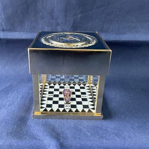 May include: A black and gold display case with a Masonic emblem on top. The case has a clear front, revealing a black and white checkered floor. A small, ornate ring with the letters "JO HT" is displayed inside.