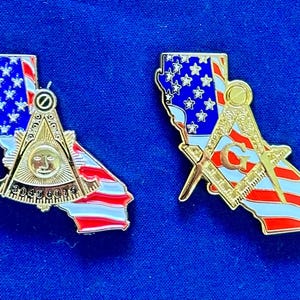 以下が含まれることがあります： カリフォルニアの形をした2つの金色のピン。アメリカ国旗のデザイン。1つのピンにはフリーメーソンのシンボルとテキスト「30456075」があります。もう1つのピンには、文字「G」が付いたフリーメーソンのシンボルがあります。どちらのピンも青色の背景にあります。