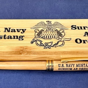 Op de afbeelding: Een houten pen en etui set. De etui is rechthoekig met afgeronde hoeken en heeft de tekst "U.S. Navy Mustang" en "Sursum AB Ordine" met een embleem. De pen is lichtbruin met gouden accenten.