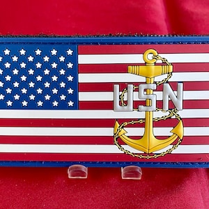 Könnte beinhalten: Rechteckiger Aufnäher mit der amerikanischen Flagge, den Buchstaben "USN" und einem goldenen Anker. Der Aufnäher hat einen blauen Rand und wird auf rotem Hintergrund angezeigt.