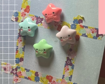 May include: Four star-shaped charms in pink, green, and white with smiling faces. The charms are arranged on a light blue background with floral patterned tape. A grid pattern and a pink rectangle are in the background.
