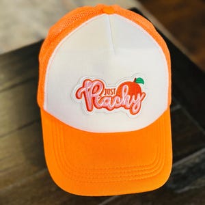 May include: Orange and white trucker hat with the text "Just Peachy" in pink script and a peach graphic. The hat features a mesh back and a curved brim, perfect for sun protection.